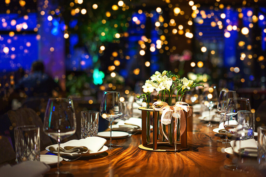 A wooden table set with white plates and napkins, clear tumblers and wine glasses, with a flower arrangement of light yellow flowers in the center, and strands of golden lights in the background